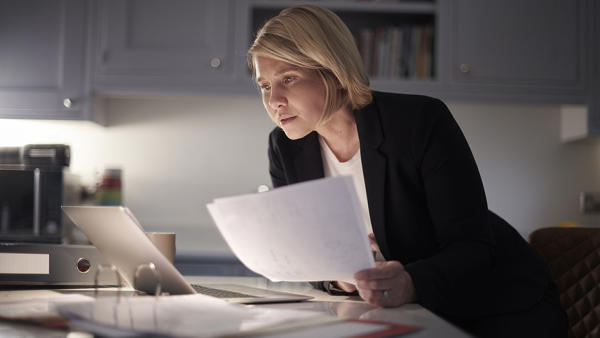 A business woman is sitting at her kitchen counter, holding up paperwork while reading from her laptop.  