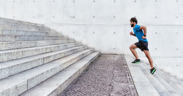 A man in a blue shirt, running shorts and bright shoes is running up wide concrete steps outdoors.  
