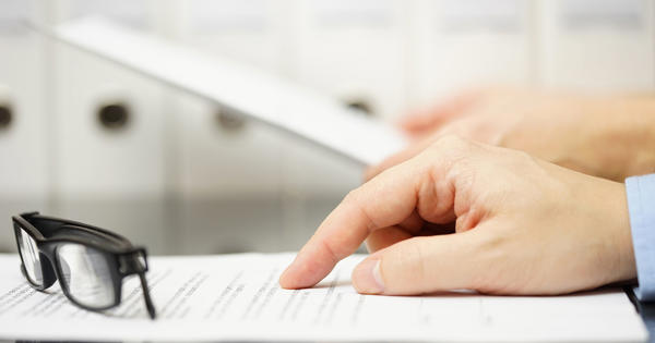 A close up photo of a man's hand pointing at a printed document with his folded glasses sitting nearby. 