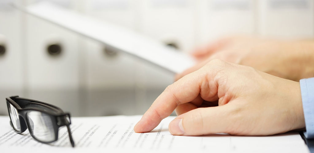 A close up photo of a man's hand pointing at a printed document with his folded glasses sitting nearby. 
